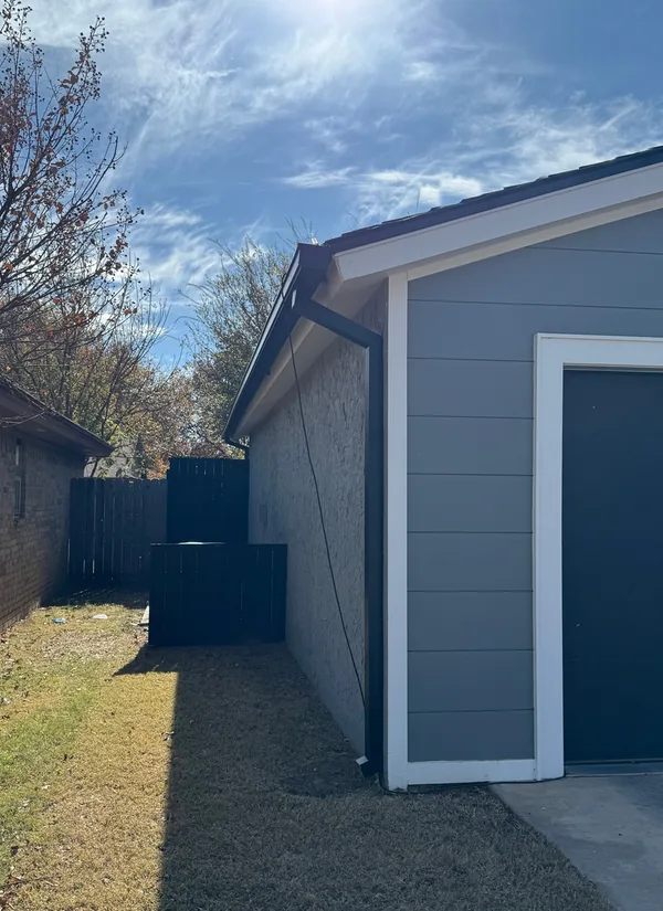 The side of a grey-sided home with a dark gutter along the roof and a downspout at the corner, under a blue sky