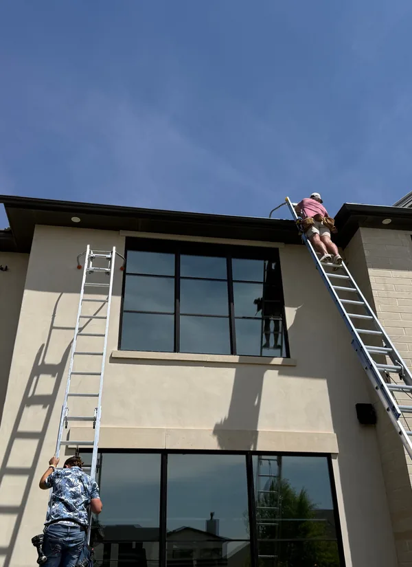 Two Premier crew members on tall ladders at the roofline of a two-storey home with large black windows, under a blue sky