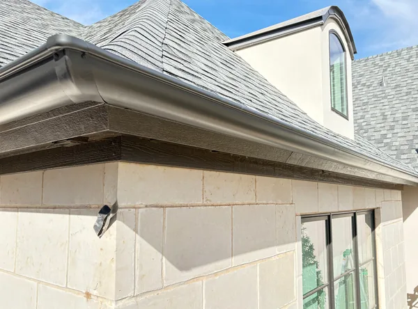 A dark seamless gutter wrapping the corner of a limestone home under a grey shingle roof
