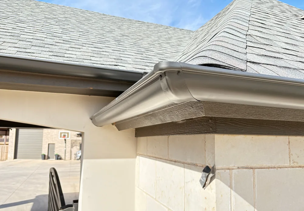 A bronze gutter on the fascia at the corner of a limestone home, the soffit under the overhang and a grey shingle roof above