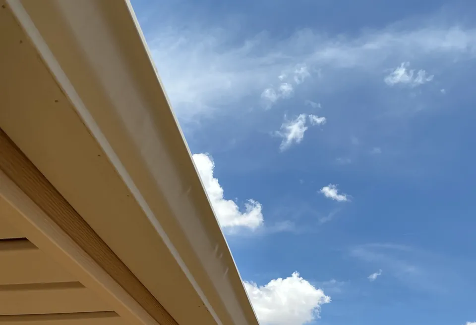 The fascia board along a roof edge with a tan gutter on it, against a blue sky.