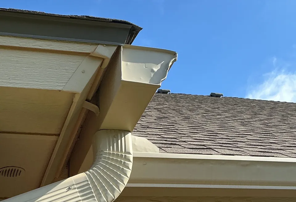 A tan gutter corner and downspout hung on the fascia, the soffit under it, against a blue sky.