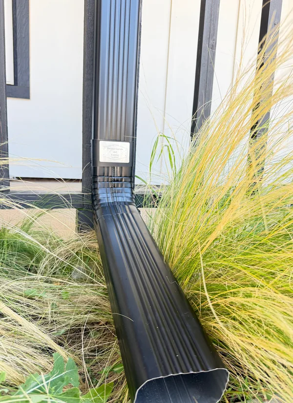 A black downspout running into a black extension through ornamental grass beside a white wall