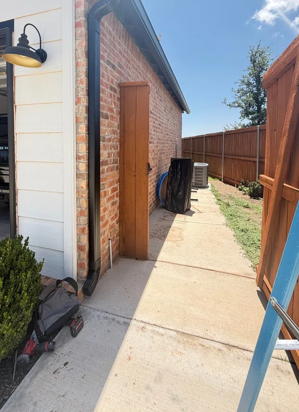 A clear concrete walk along the side of a white-sided and brick home, a ladder at the fence and a black downspout down the corner