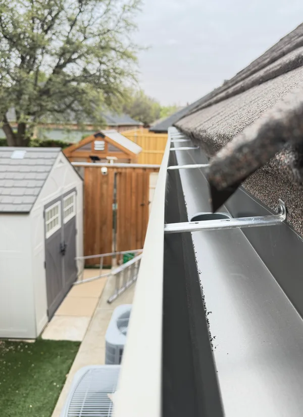 Looking along the inside of a clean cream gutter run on a shingle roof edge, its hangers spaced along it, a shed and a fence beyond