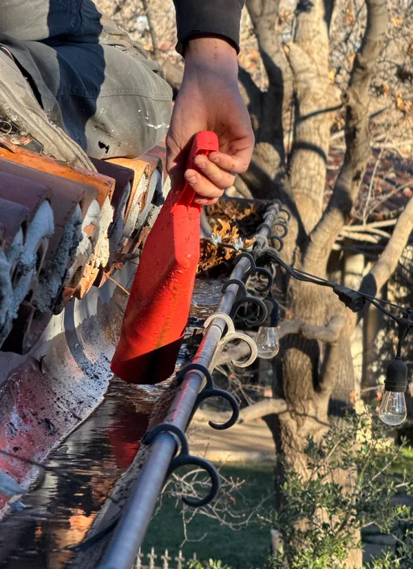 A crew member’s hand in a red glove scooping leaves out of a half-round gutter along a clay tile roof