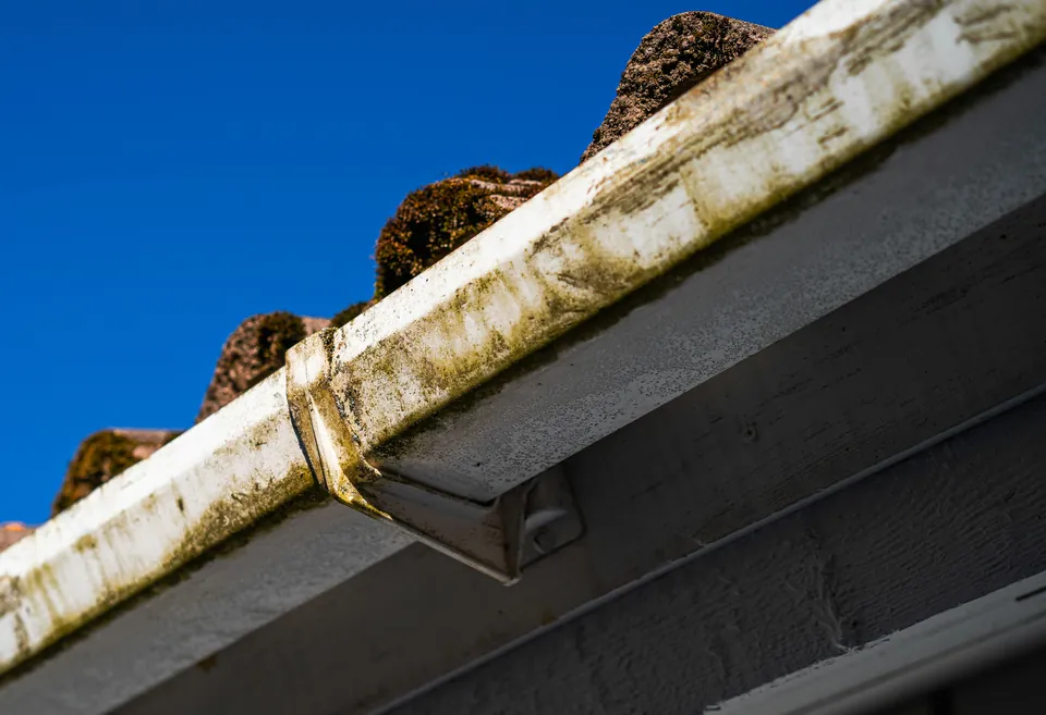 A white gutter along a roof edge, its front face streaked with dirt and algae, against a deep blue sky.