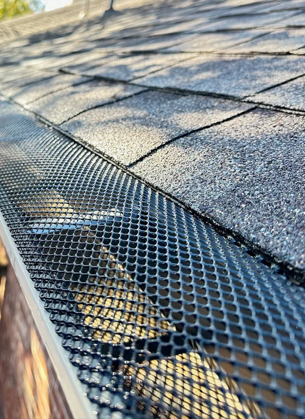 A black mesh gutter guard fitted on a gutter under gray shingles.
