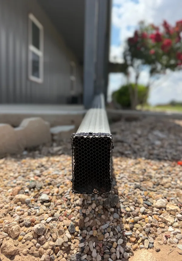 A black mesh guard fitted in a downspout's outlet over gravel, grey siding behind