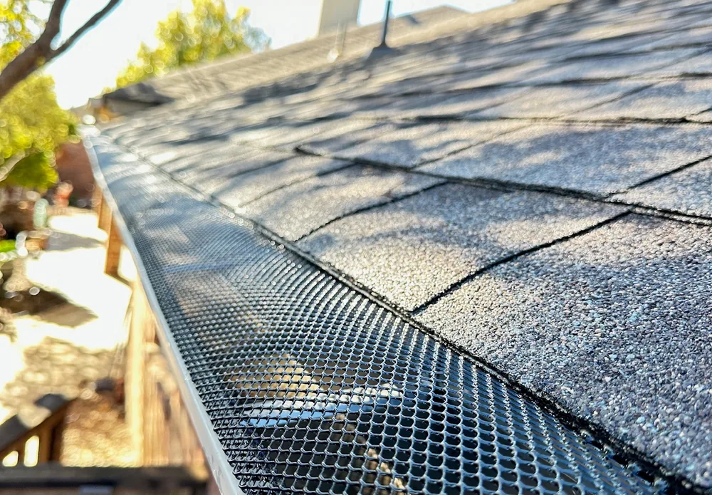A black mesh gutter guard fitted along a gutter under grey shingles, a tree against the sky behind