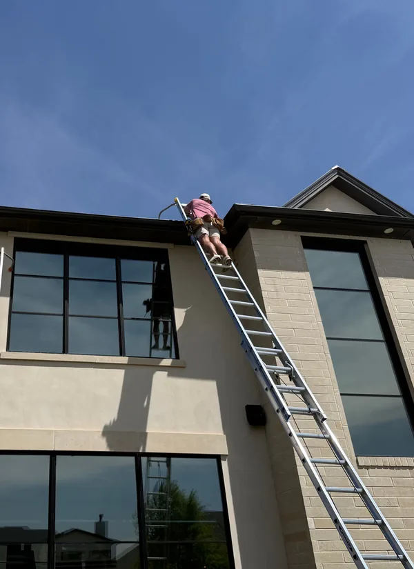 A Premier crew member at the top of a tall ladder at the roofline of a two-storey home with large black windows, under a blue sky