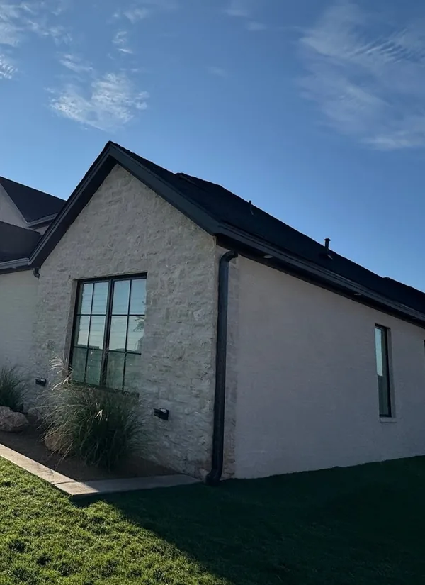 A limestone and stucco home with a gable, dark gutters along its rooflines and a clear lawn, under a deep blue sky