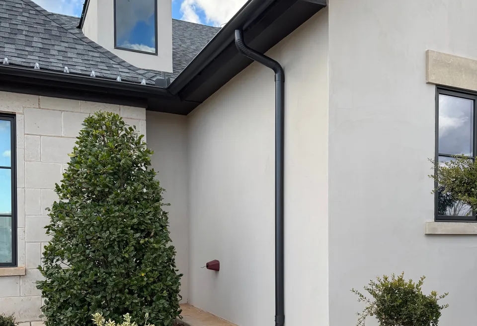 A black gutter along the roof edge of a white stucco home under a dormer, its downspout running down the wall, against a blue sky.