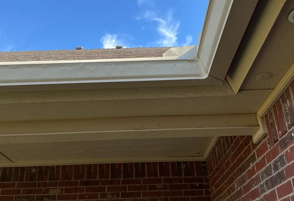 A tan gutter along the roof edge of a brick home, seen from below, with the fascia board and the soffit under it against a blue sky.