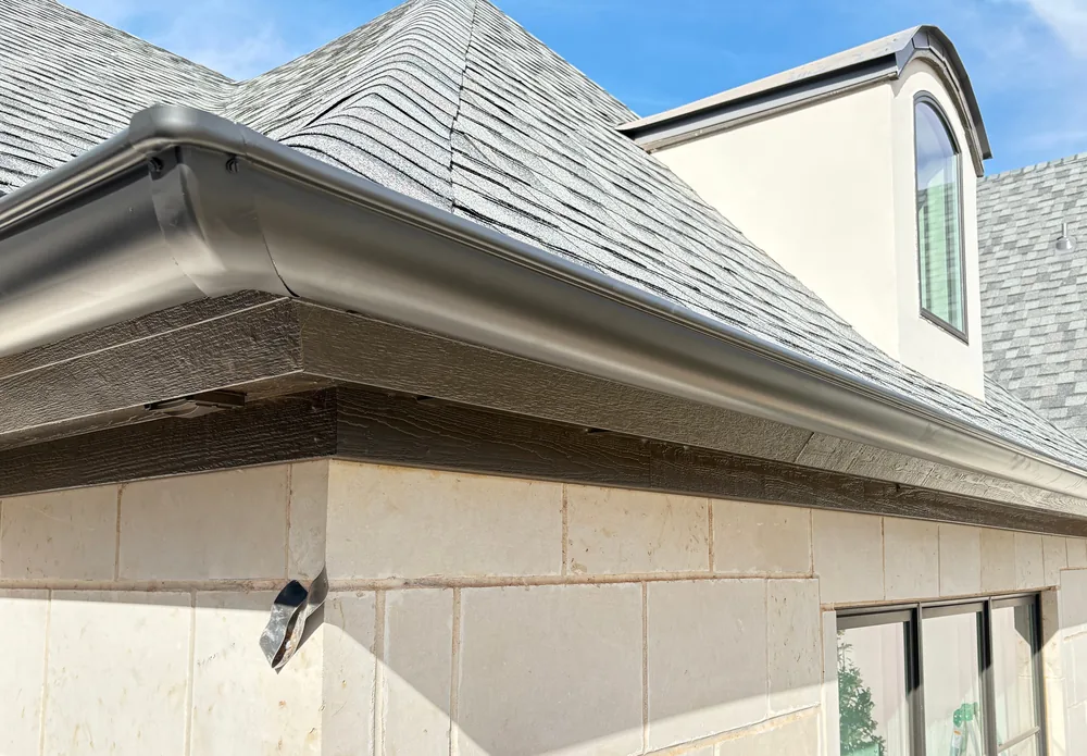 A dark bronze seamless gutter running along the eave of a limestone home and round its corner, a dormer behind, under a blue sky
