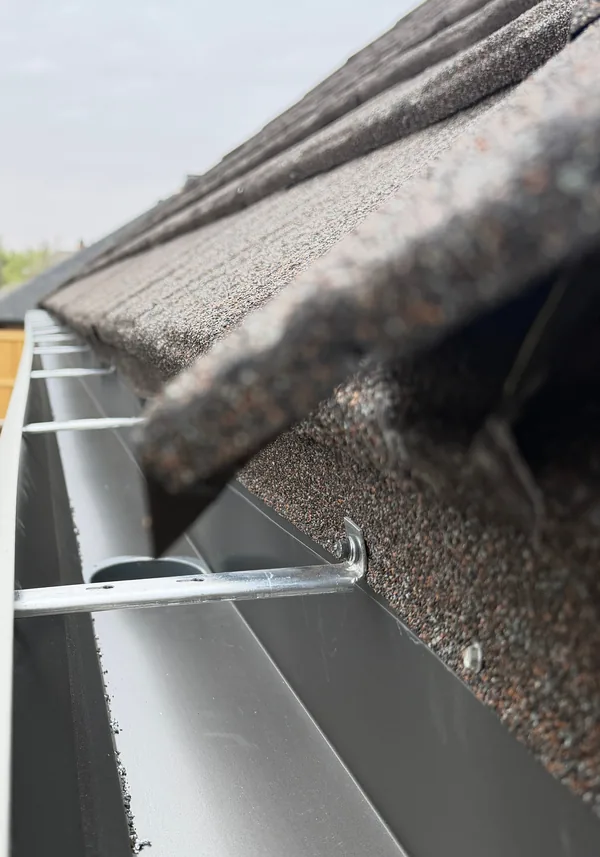 Looking along the inside of a new aluminum gutter under a shingle roof, its hangers spaced along the run