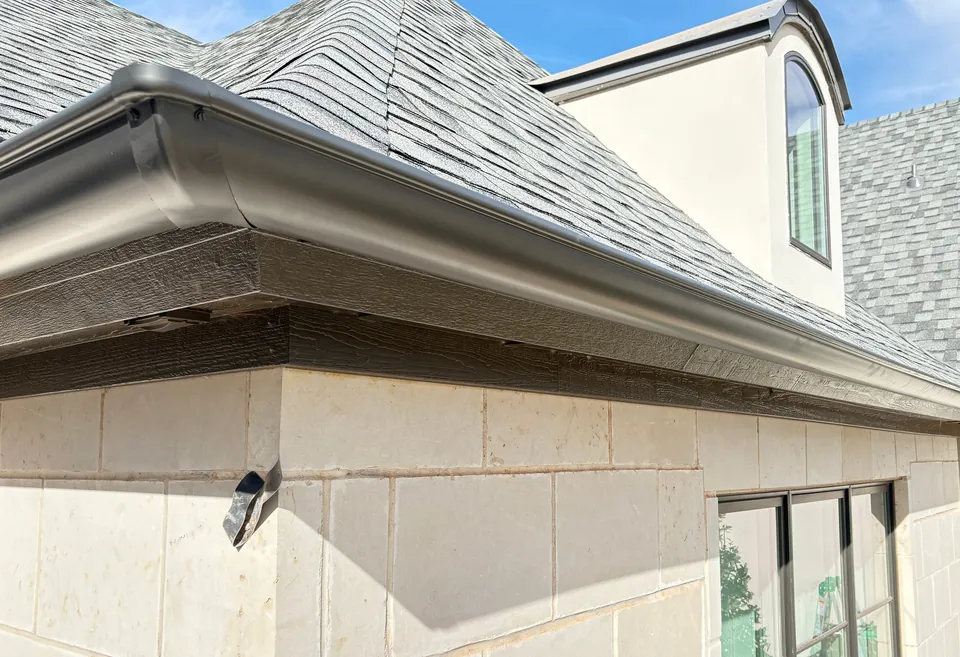 A new dark bronze seamless gutter wrapping a limestone corner under a gray shingle hip roof and a dormer.
