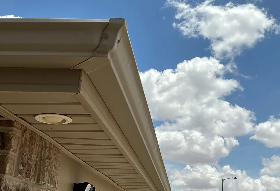A tan gutter on the fascia at a roof corner, the soffit under it, against a cloudy blue sky.