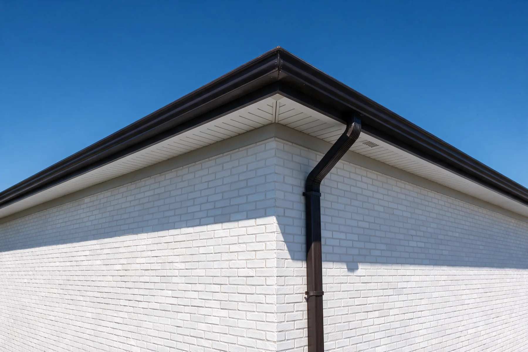 A new dark bronze seamless gutter and downspout at the corner of a white painted brick house under a clear blue sky