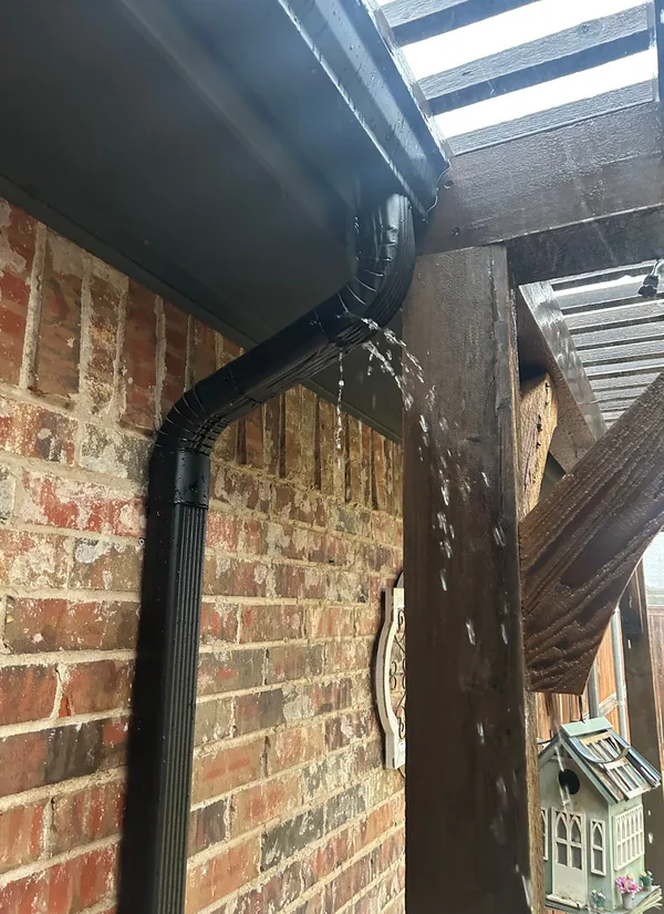 Water spraying from the joint where a black gutter outlet meets its downspout elbow, on a brick wall beside a wooden pergola post.
