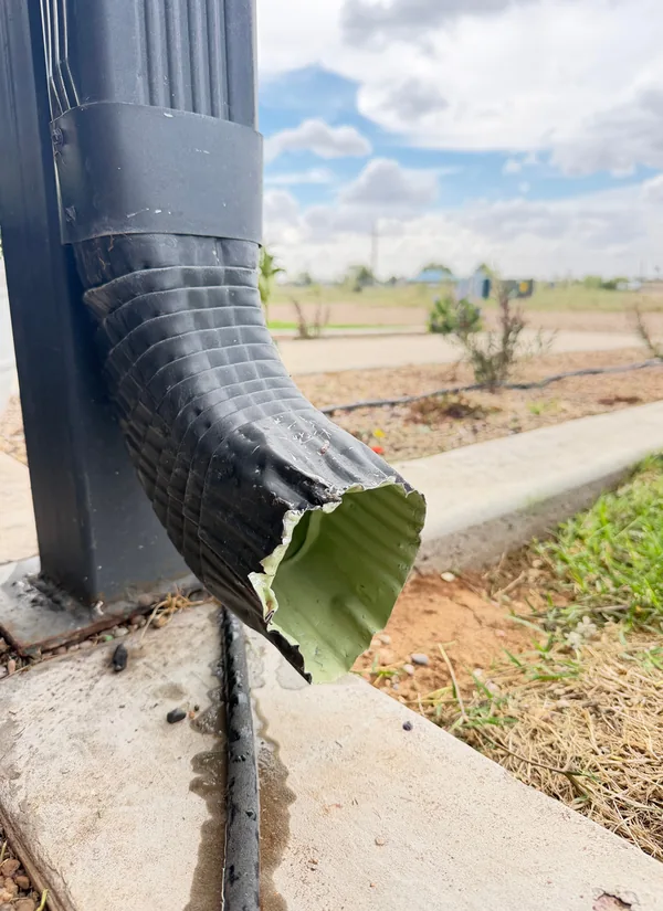 A crushed, torn downspout outlet at the edge of a concrete slab, under a blue sky with clouds.