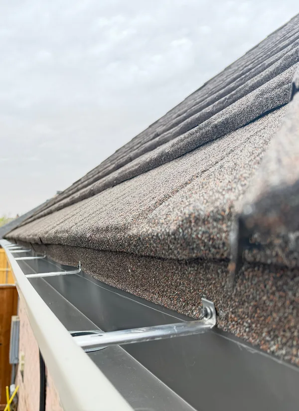 Looking along the inside of a cream gutter on a shingle roof edge, with a metal hanger across the trough.