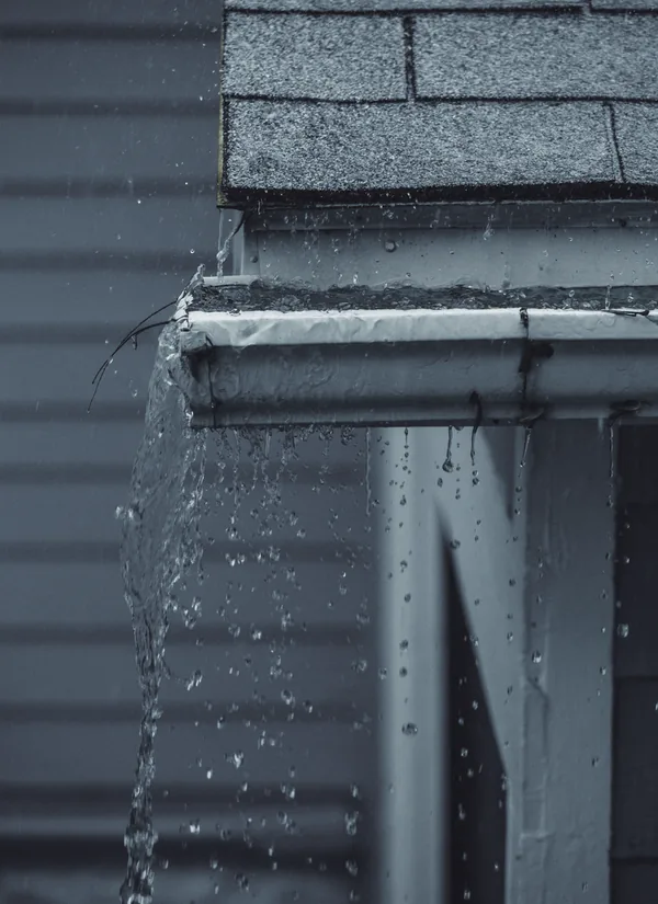 Rain pouring over the end of a white gutter on a house with teal siding, the water falling in a sheet and in drops down the wall.