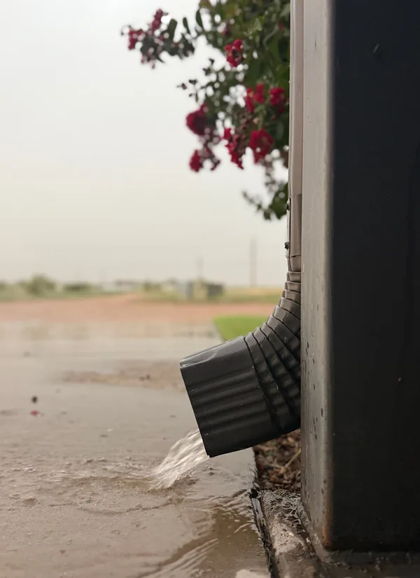 A black downspout outlet pouring rainwater onto a flooded yard