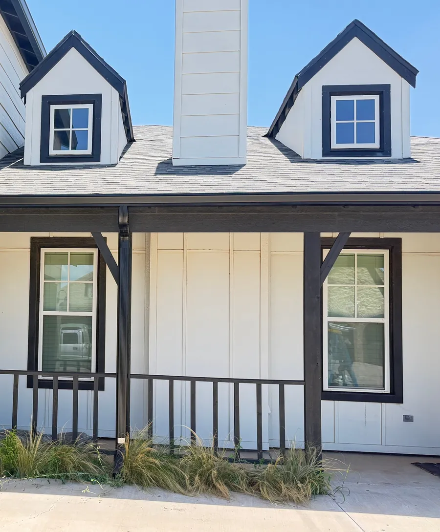 A white farmhouse with a chimney between two dormers, a covered porch and black gutters, under a clear blue sky.