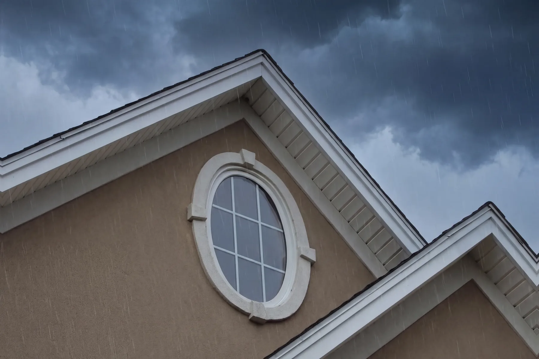 An oval window with white trim in the tan stucco gable of a home, in the rain under dark storm clouds
