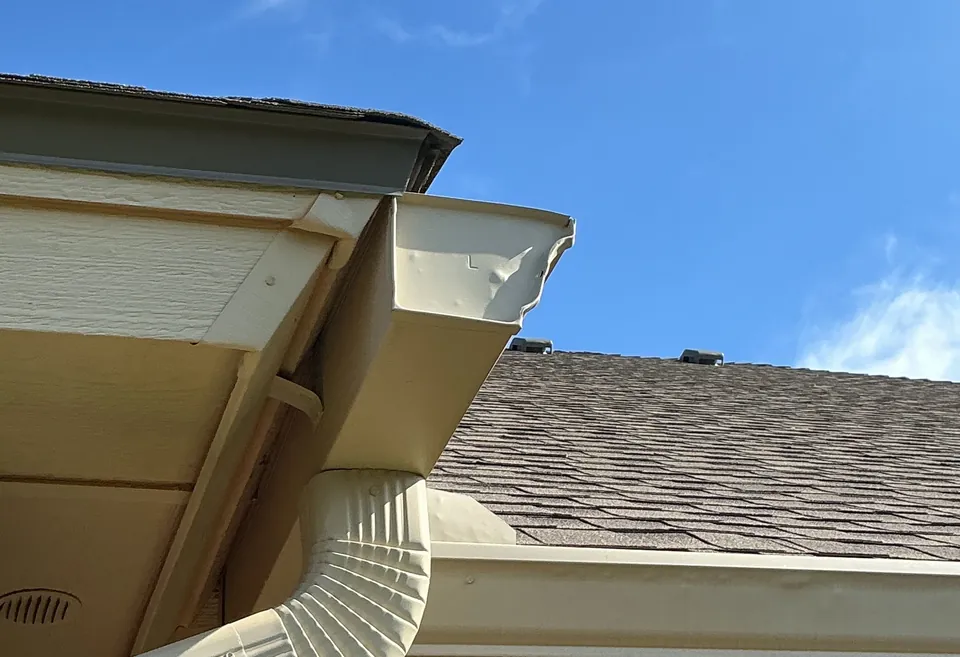 A tan gutter corner and downspout under the soffit and fascia, against a blue sky.