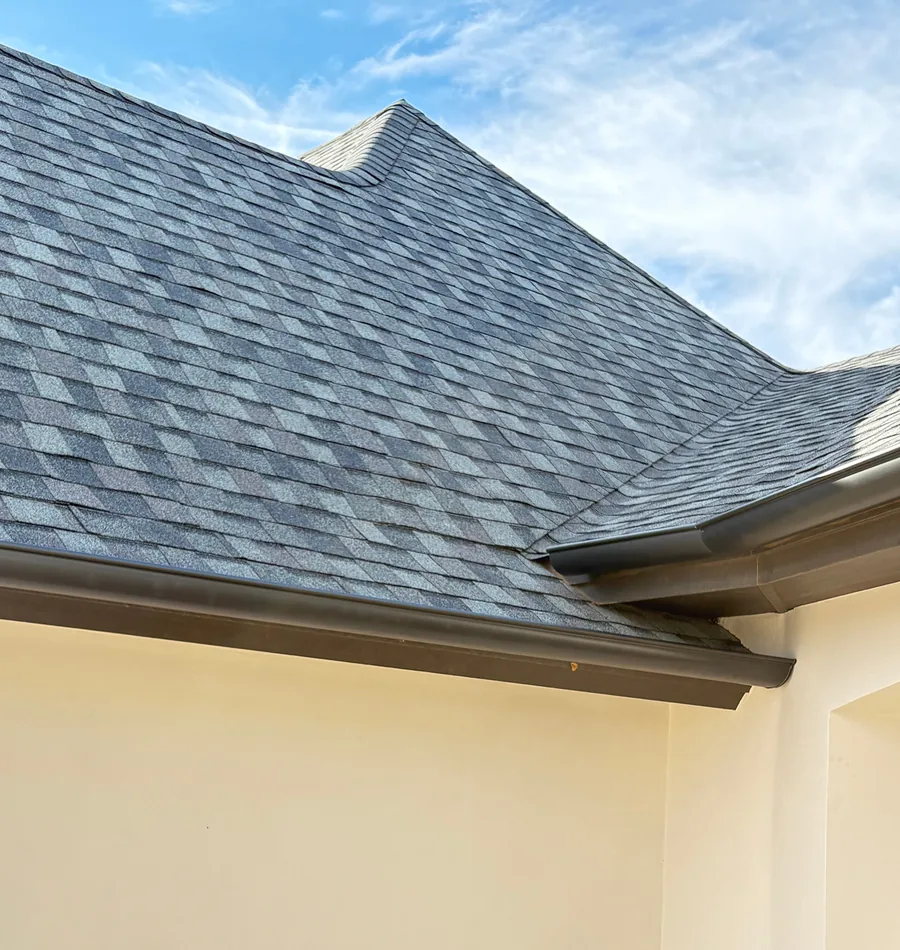 Dark gutters along the eaves of a white stucco home where two grey shingle roofs meet, under a blue sky.