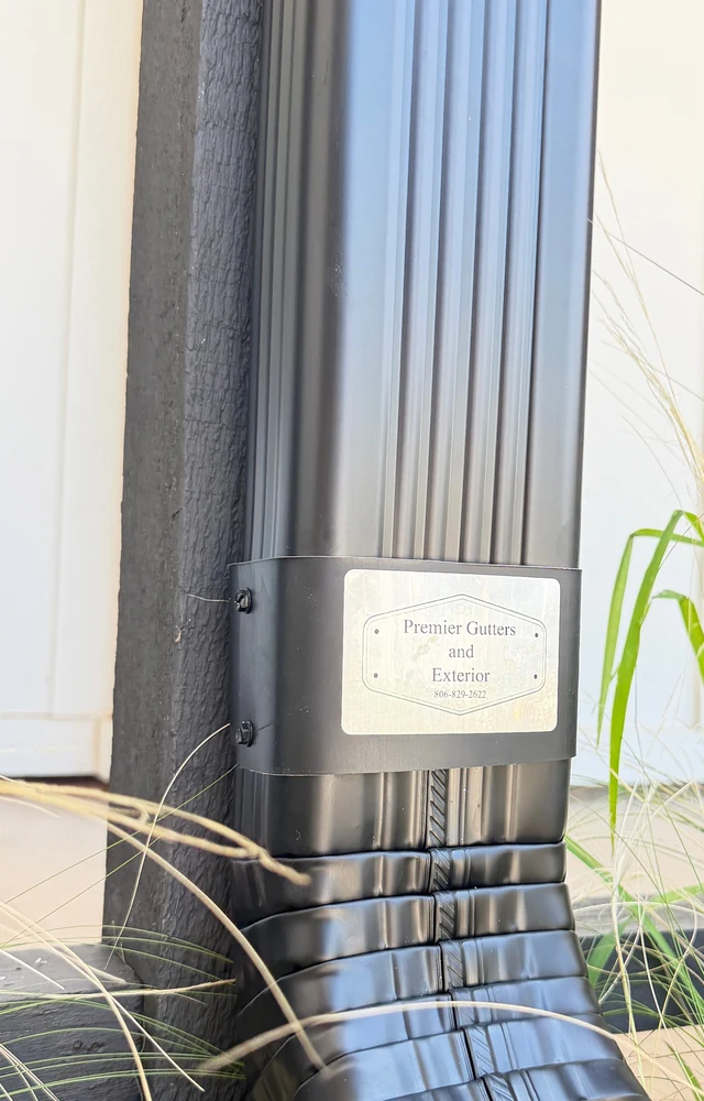 A black fluted downspout with a Premier Gutters and Exterior nameplate fixed to it, against a white board-and-batten wall and a black post, near Lubbock.