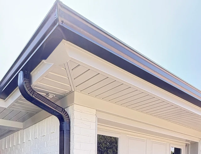 A dark bronze gutter wrapping a roof corner above a white soffit, and its downspout curving down a white painted brick wall beside a garage door, near Lubbock.