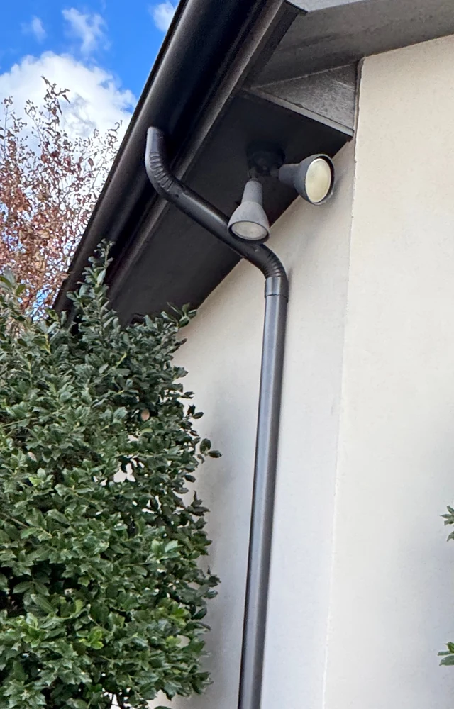 A dark gutter and its black downspout curving down a white stucco wall past an outdoor light, beside a green shrub, under a blue sky near Lubbock.
