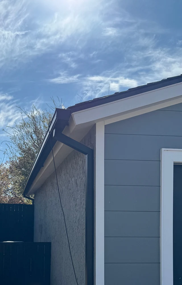 A dark gutter and downspout at the corner of a gray-sided home with white trim and a garage door, under a blue sky with thin clouds near Lubbock.