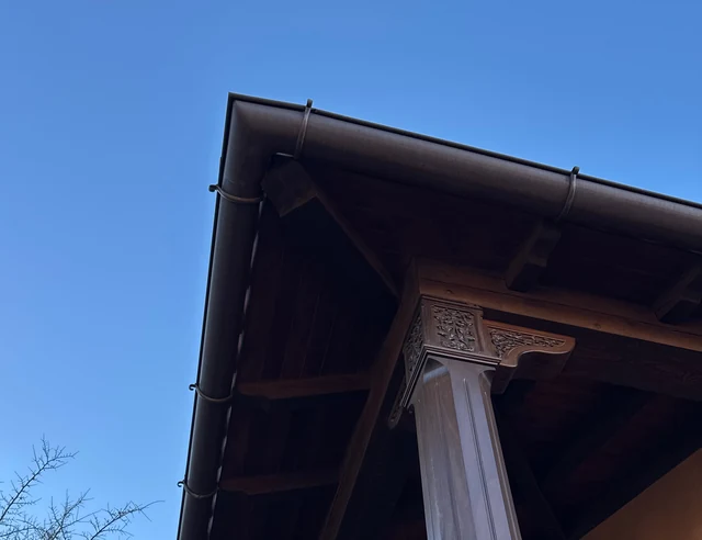 A dark bronze half-round gutter wrapping the corner of a carved wood porch roof against a deep blue sky, near Lubbock.