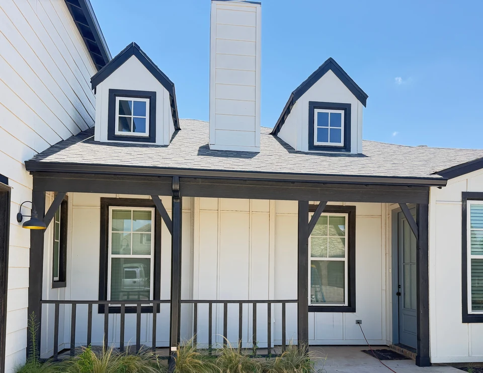 A white board-and-batten farmhouse with black trim, two dormers, a chimney and dark gutters along the porch roof, under a blue sky near Lubbock.