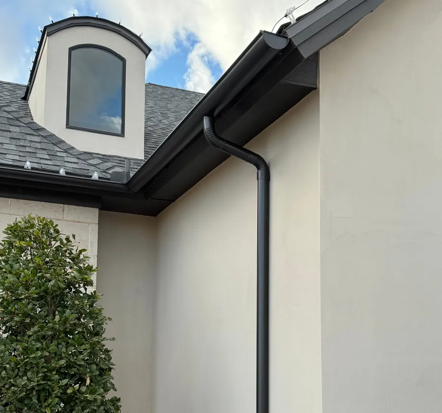 A black gutter and downspout on a white stucco home under a dormer and a blue sky.