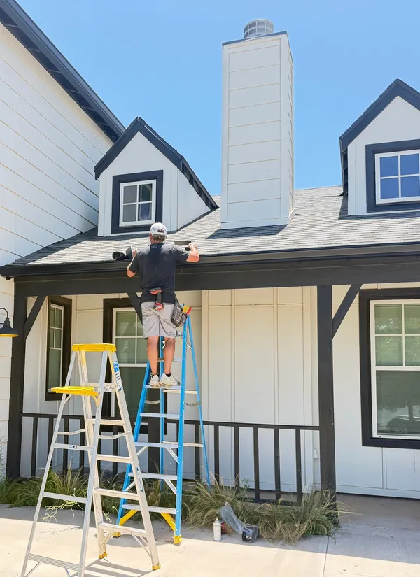 A crew member on a ladder at the porch roof of a white farmhouse.