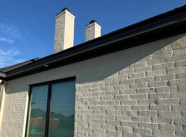 A black seamless gutter along the eave of a grey painted brick home, under two white chimneys and a blue sky