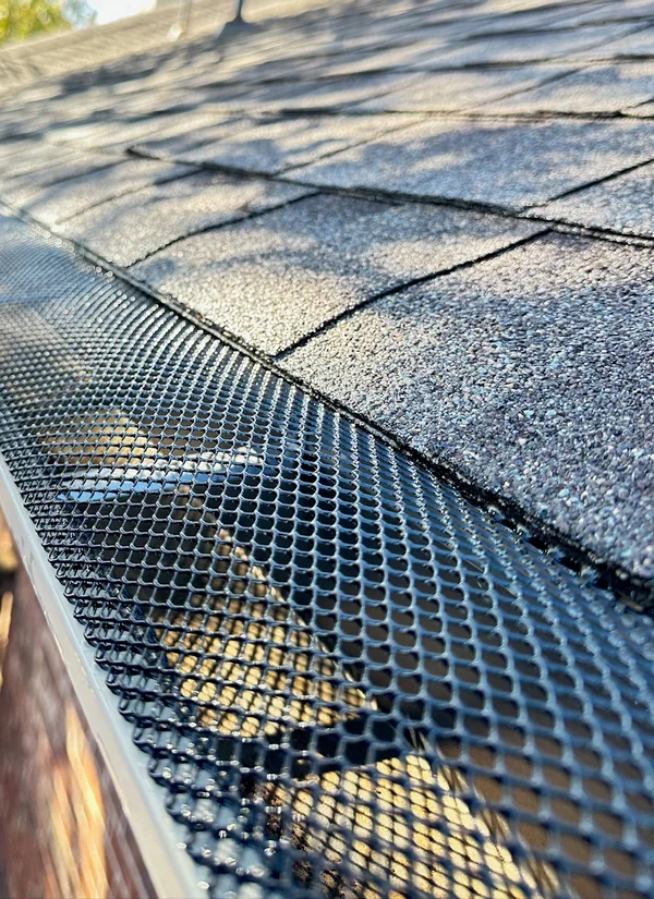 A black mesh gutter guard fitted over a gutter along the edge of a gray shingle roof.