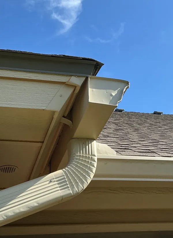 A tan gutter corner and downspout under the soffit and fascia, against a blue sky.