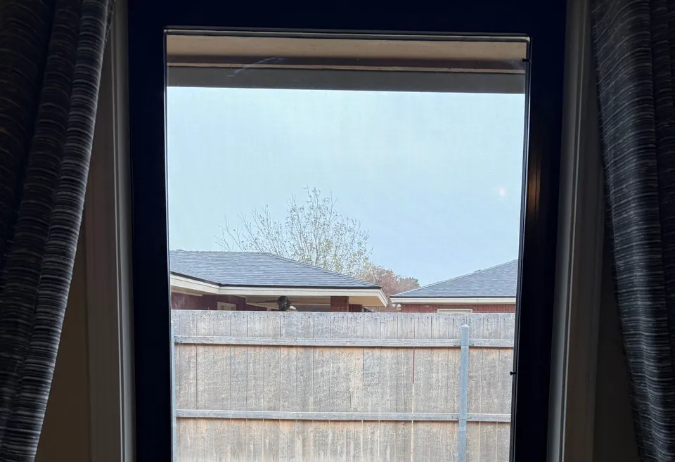 A black-framed window seen from inside between gray curtains, its clear glass looking onto a fenced yard.