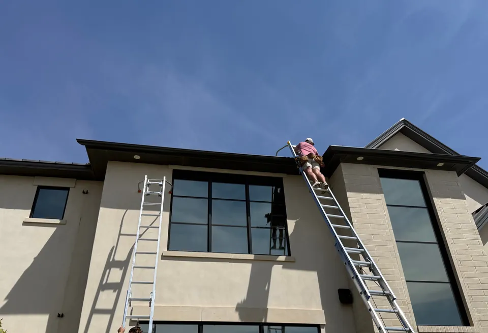 Two crew members on tall ladders at the big black windows of a two-storey stucco and stone home, under a deep blue sky.
