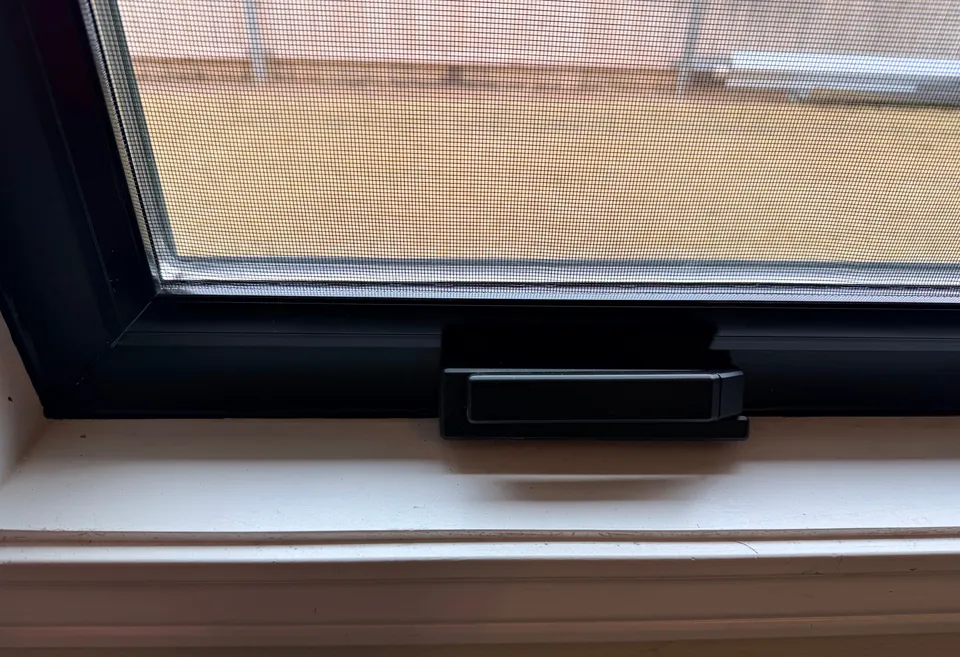 A close look at a black window frame and its latch, the screen mesh across the glass, above a white sill.