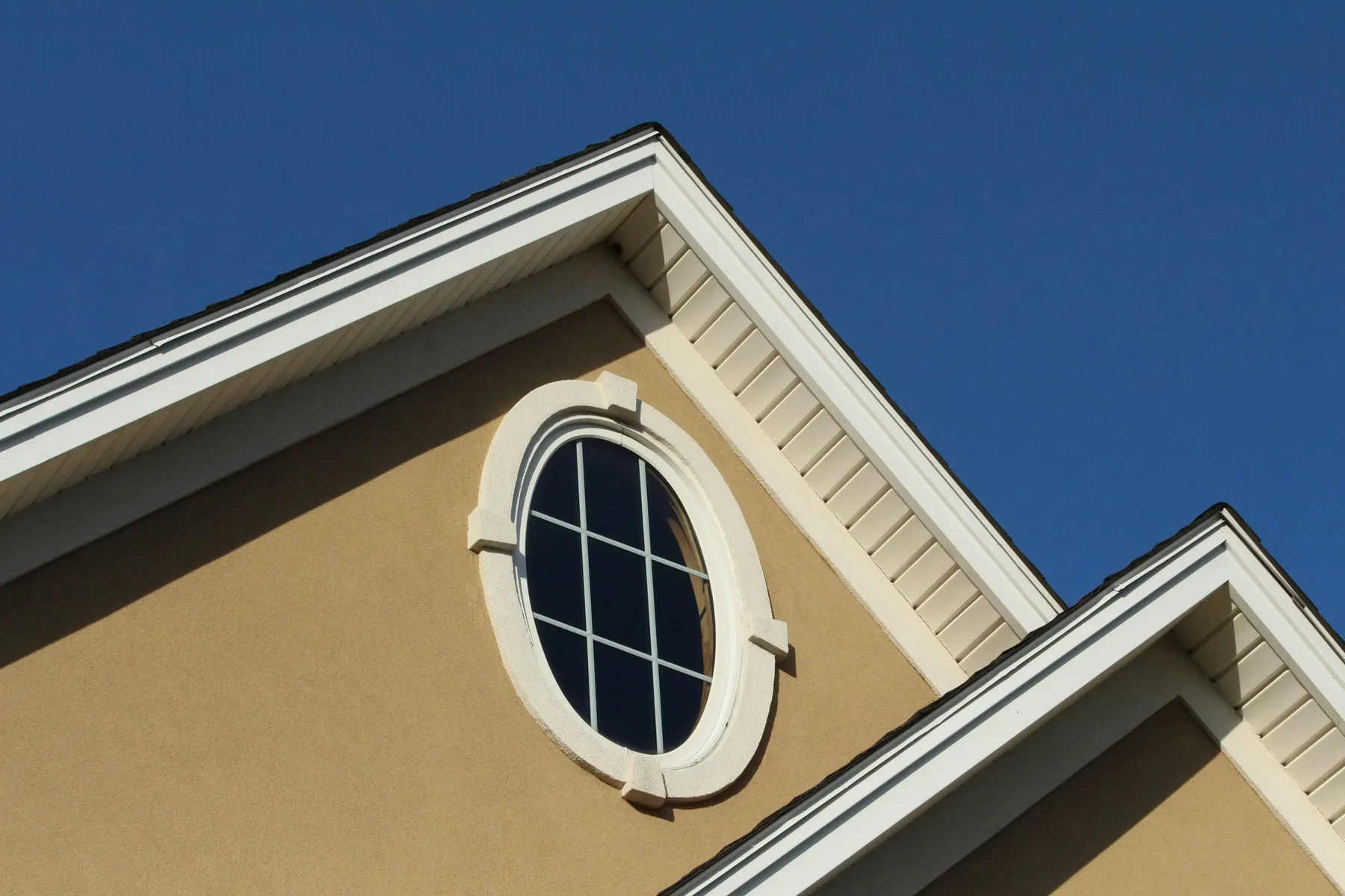 An oval window with white trim in the tan stucco gable of a home, under a deep blue sky