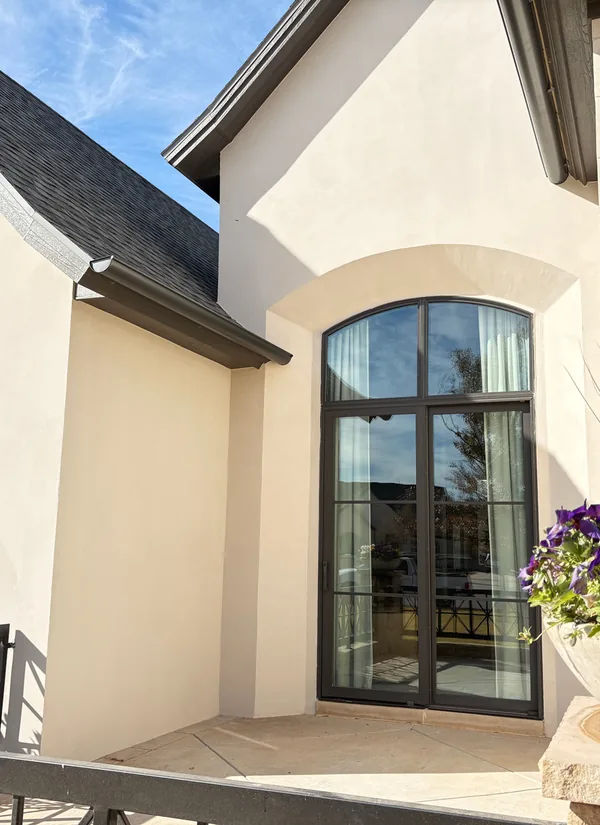 A cream stucco home with a tall black arched window under its gable and a grey shingle roof, under a blue sky