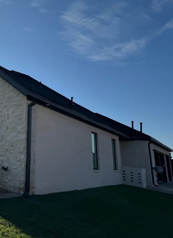 The side of a limestone home with black windows and a dark gutter along its roof, a green lawn, under a deep blue sky